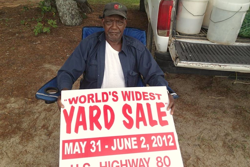 A man sits with a sign from the first "World's Widest Yard Sale" in 2012. 