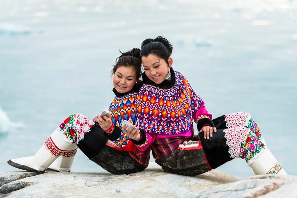 Two girls wear Greenland's national costume while taking photos on their iPhones. 
