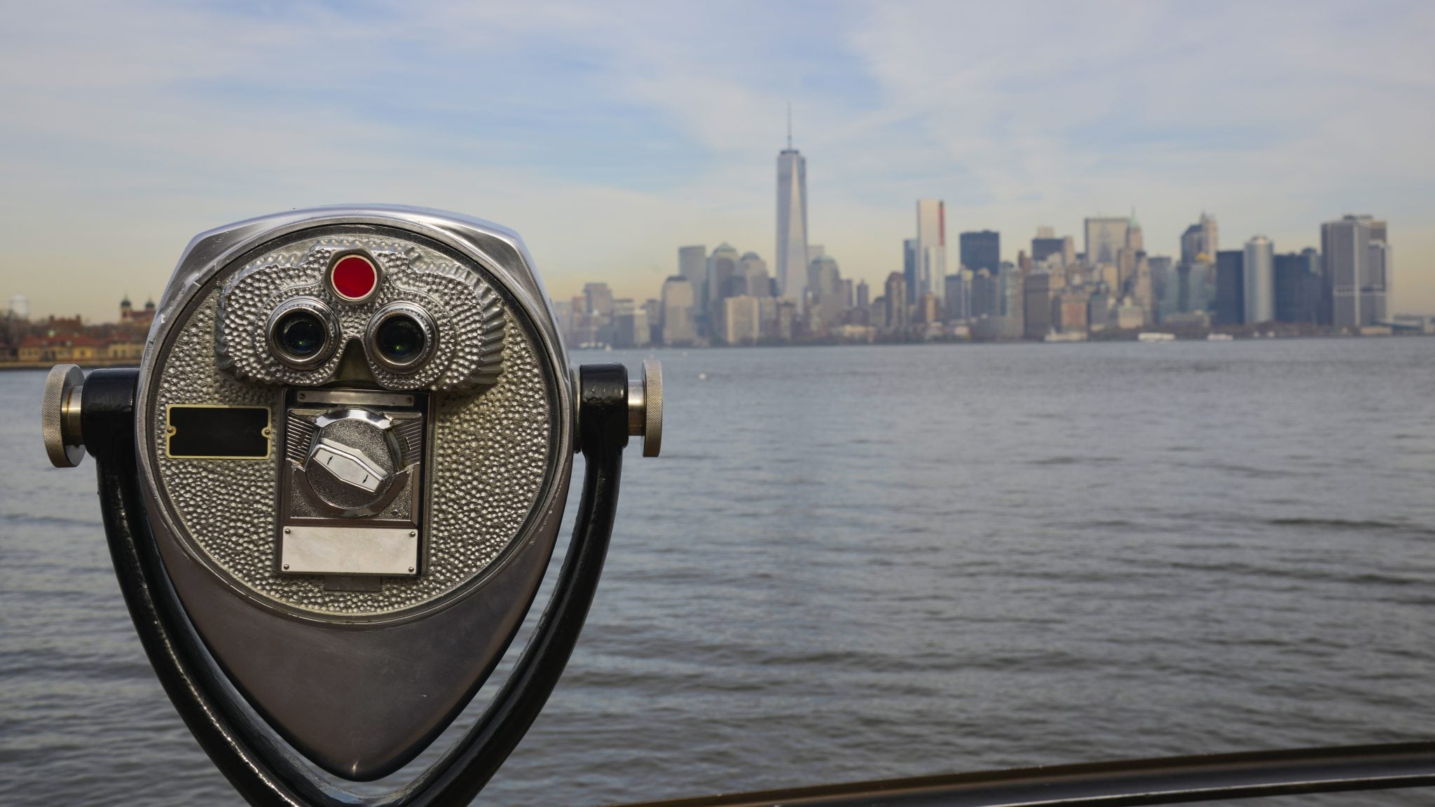 A tourist telescope on the Ellis Island and New York City Skyline with the Hudson River and the Lower Manhattan skyscrapers in the background on a sunny summer day.