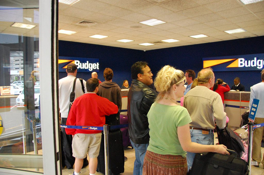 Travelers are waiting on a long line in a crowded Budget car rentals site. Booking sites have hopes to make this process quicker with connected trips.