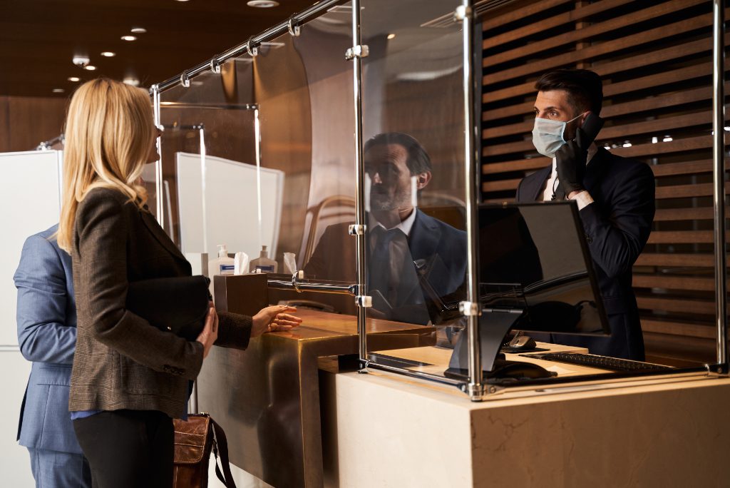 Male receptionist wearing a medical mask and rubber gloves while being at work and looking at the visitors with a phone in his hand. 