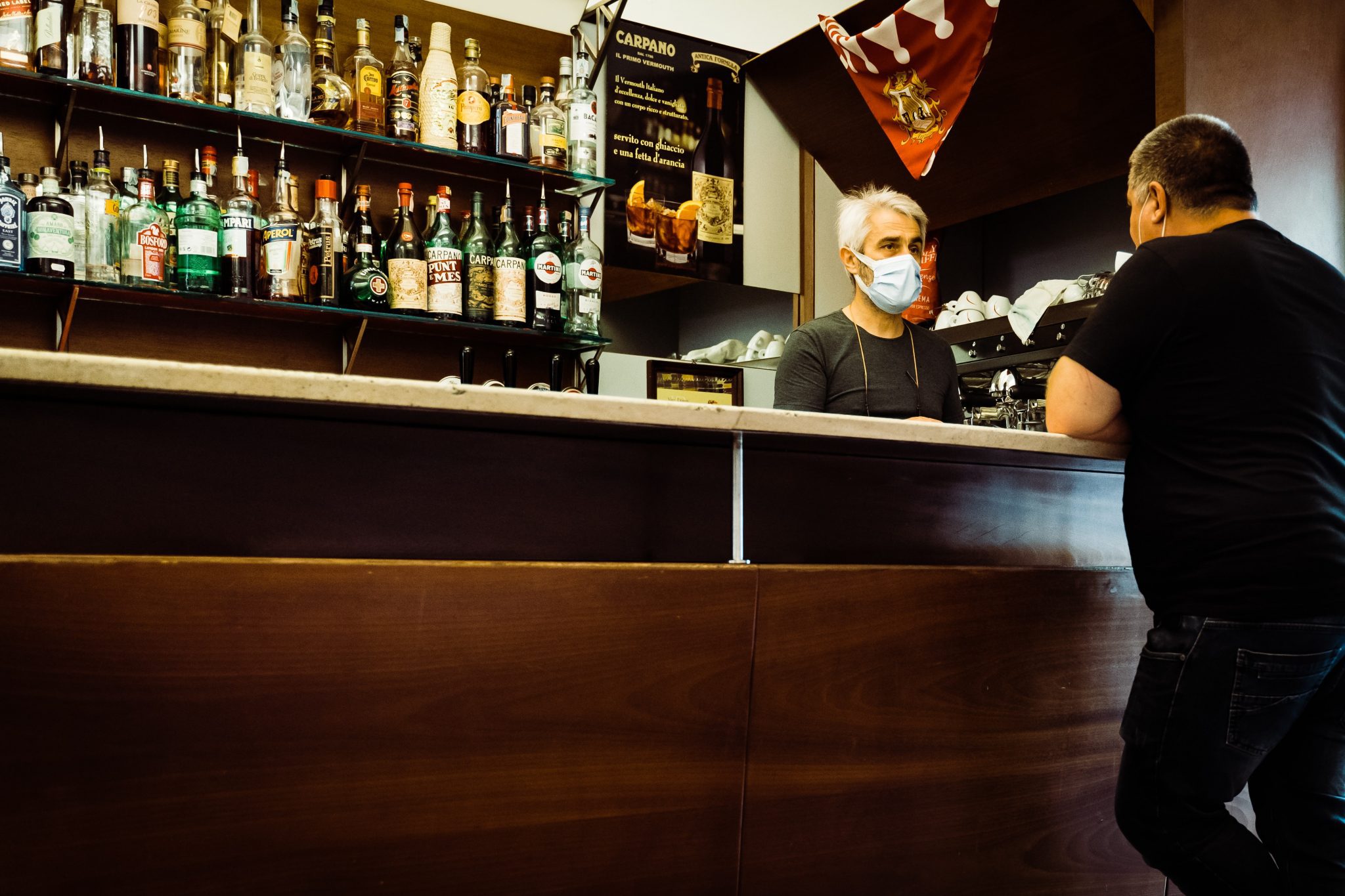 A bartender wearing a face mask chats with a customer in a coffee shop in Tuscany, Italy.  