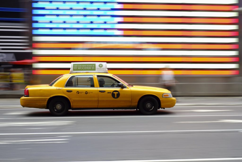 A New York City yellow cab drives by an American flag. Credit: Flickr.com/Nick Harris https://flic.kr/p/dd3KAh 