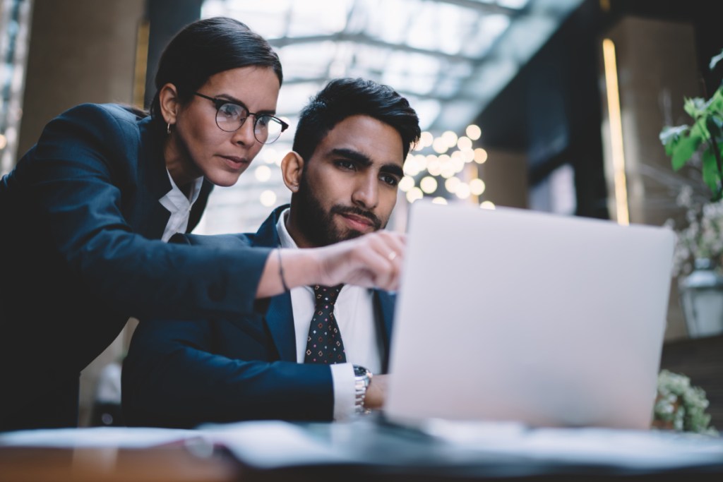 Focused businesswoman showing information on laptop screen to focused male colleague sitting at table with documents in lobby of hotel