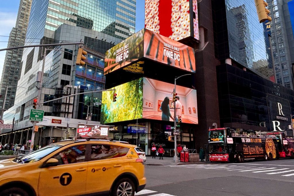 A street scene with a speeding taxi, tourist bus, and large video advertisements rising into the sky