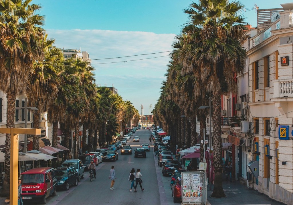 A street lined with buildings, trees, and cars. Pedestrians walk in the road.