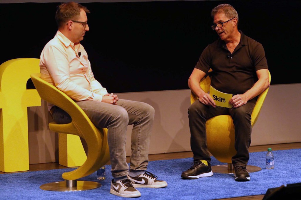 Two men on stage talking, sitting by yellow "Skift" sign