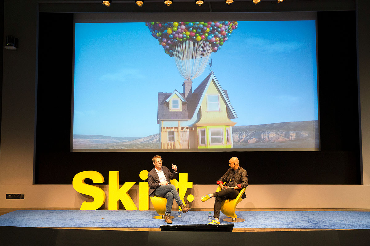 two men speaking on stage with a screen showing a floating house behind them.