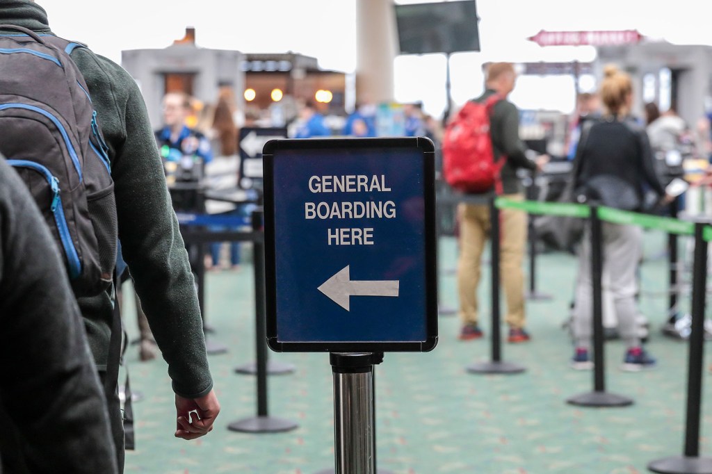 passengers lined up at airport security to the left of a sign showing them what to do.