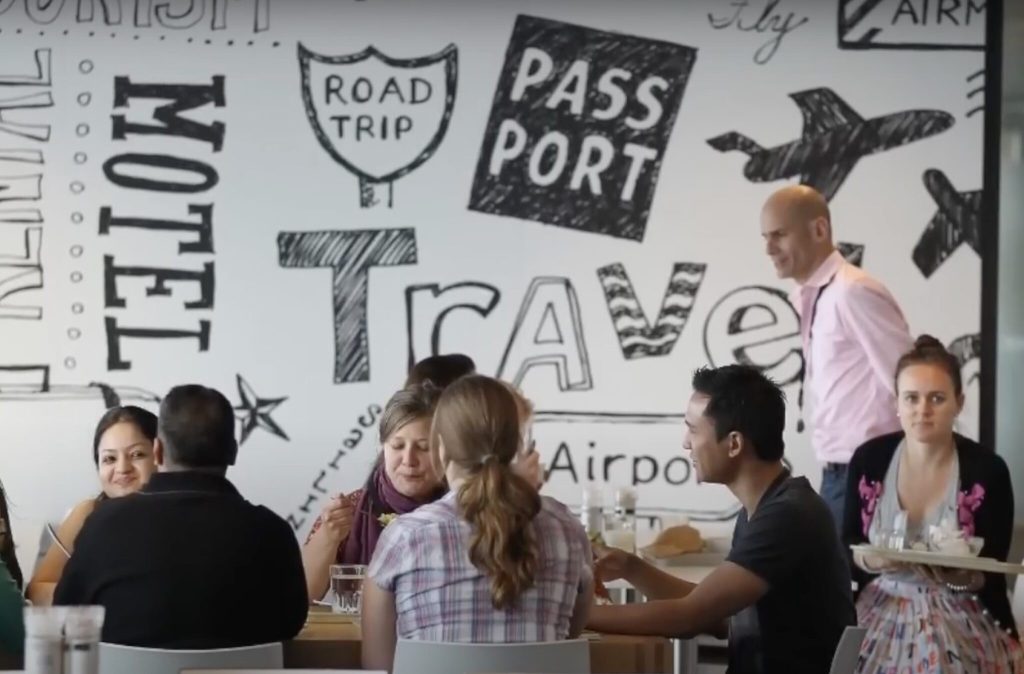 Employees sitting at a lunch table with travel words like "passport" on a whiteboard behind them.