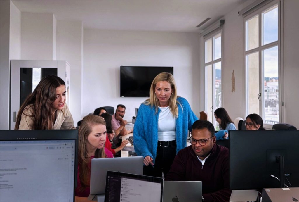 Employees in an office looking at some computers. Other employees sit at a table in the background.