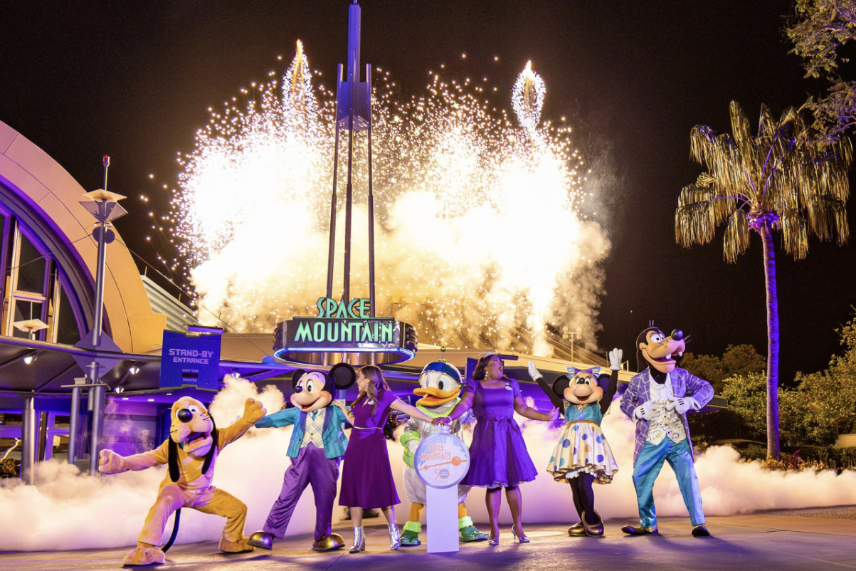 Space Mountain is light up at night with Mickey Mouse and other characters standing outside it.