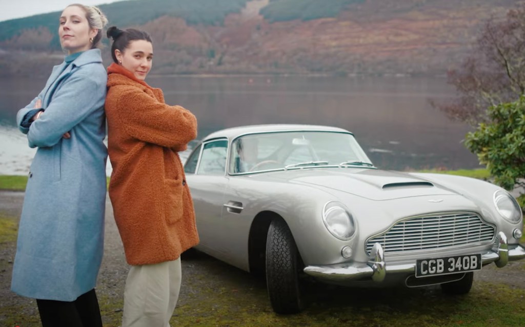 2 women stand next to a car with a scenic view in the background.