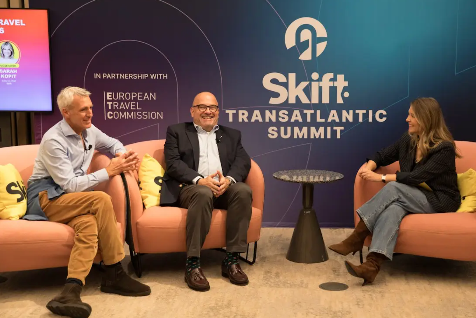Three people sit and converse on stage at the Skift Transatlantic Summit, with event branding and sponsor logos visible in the background.