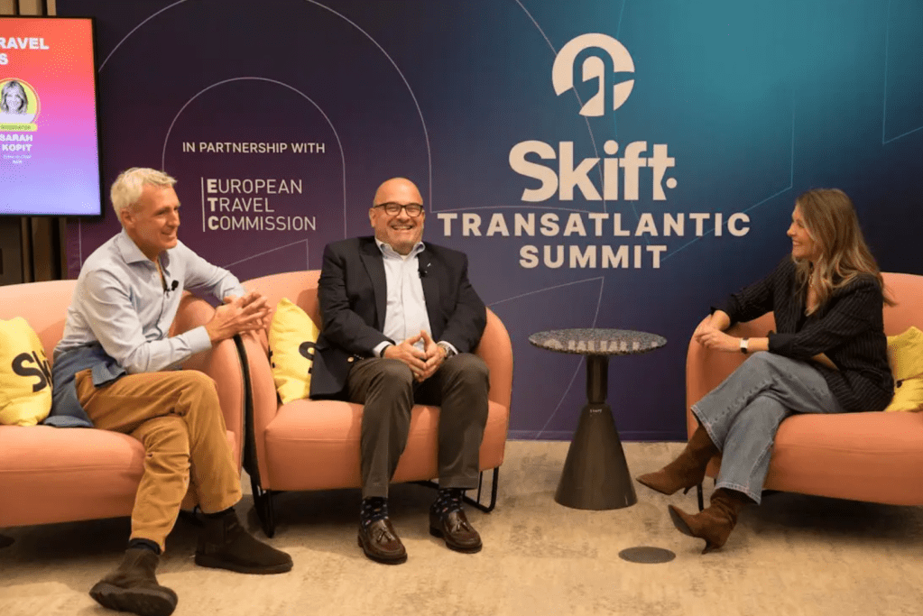 Three people sit and converse on stage at the Skift Transatlantic Summit, with event branding and sponsor logos visible in the background.